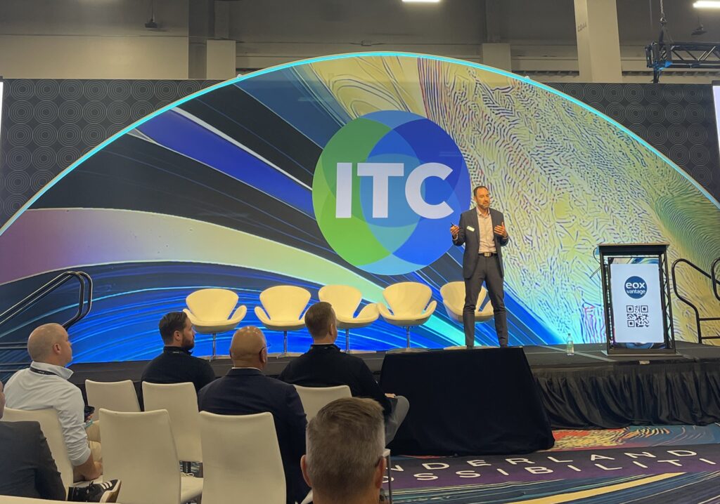 Travis Roe standing on a stage in front of a small audience in front of a colorful backdrop at ITC 2025.