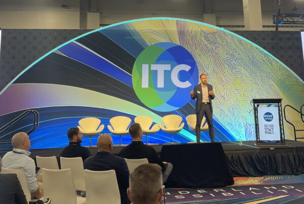 Travis Roe standing on a stage in front of a small audience in front of a colorful backdrop at ITC 2025.