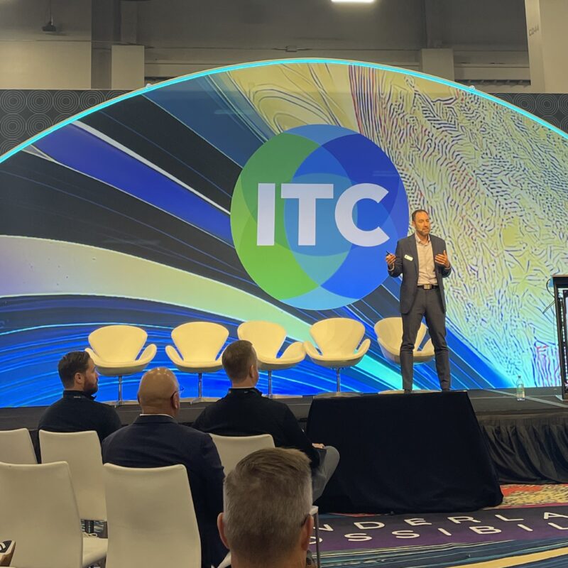 Travis Roe standing on a stage in front of a small audience in front of a colorful backdrop at ITC 2025.